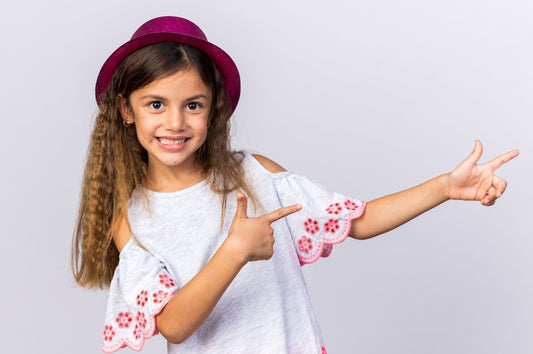 smiling-little-caucasian-girl-with-purple-party-hat-pointing-side-isolated-white-wall-with-copy-space